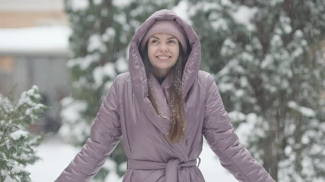 Joyful slim young woman tossing snow outdoors smiling. Portrait of beautiful Caucasian brunette lady having fun on snowy winter day outdoors. Happiness and leisure concept