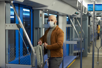 A bald man with a beard in a face mask is putting on a backpack in a subway car.