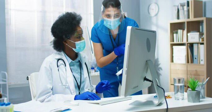 Work in hospital. Coronavirus pandemic. Caucasian young beautiful female nurse talking to African American woman doctor holding documents analysis results and looking at computer screen in cabinet