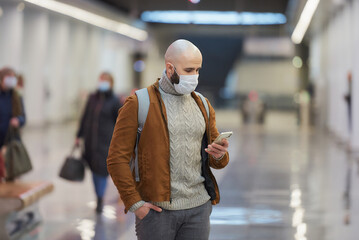 A man in a face mask is using a smartphone while waiting for a subway train.