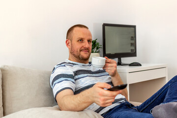 Fototapeta na wymiar Shot of a handsome young man watching television at home. Happy young Caucasian man holding cup of coffee and remote controller while sitting on sofa at home. Leisure, technology and people concept.
