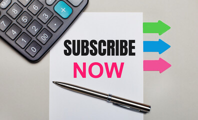 On a light gray background, a calculator, a white sheet with the text SUBSCRIBE NOW, a pen and bright multicolored stickers. View from above