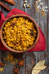 Cooked spicy rice in a wooden bowl and various spices on a dark background. Rice with cumin, turmeric, star anise and other spices. Indian food.