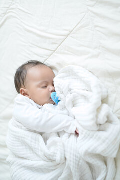A recently born half-born baby is wrapped in a white cloth and slept in bed while sucking on a rubber nipple