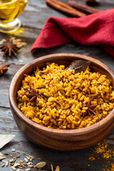 Cooked spicy rice in a wooden bowl and various spices on a dark background. Rice with cumin, turmeric, star anise and other spices. Indian food.
