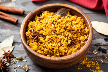 Cooked spicy rice in a wooden bowl and various spices on a dark background. Rice with cumin, turmeric, star anise and other spices. Indian food.