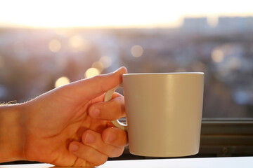 Fresh start in the morning, male hand with cup on sunrise background. Cozy atmosphere, city view from the window