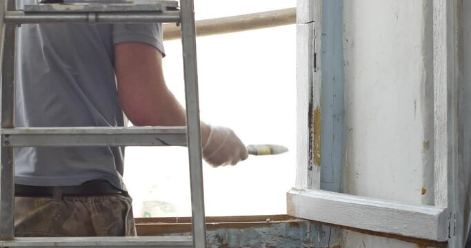 Handyman paints a window frame with white paint with a paint brush. Hand in latex rubber gloves applies a glossy paint finish to window casement. Repairman painting old window at table indoors