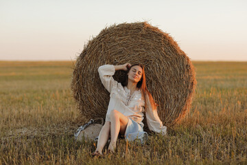 Naklejka na ściany i meble young woman in the beautiful light of the summer sunset in a field is sitting near the straw bales. beautiful romantic girl with long hair outdoors in field