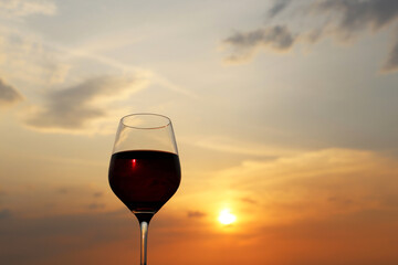 Glass with red wine on sunset background, evening sun and sky with clouds reflected in the glass. Concept of celebration, wine industry