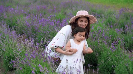 Motion of young pretty mother dancing with little daughter in lavender field, sitting on ground. Loving smiling woman holding kid hands, having fun, hugging and kissing. Concept of motherhood, nature. - Powered by Adobe