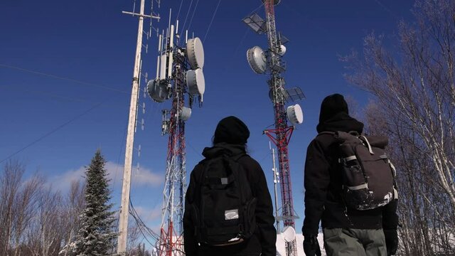 Locked down shot of two tourists walking up to a cellular transmission base on a snowy hill, and excitedly waving at camera at the end.