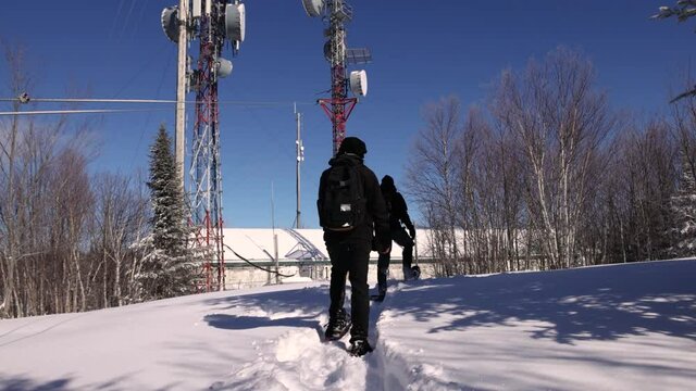 Videography of two explorers walking up the snowy mountain, at the base station of 4G cellular network towers, surrounded by bare trees.