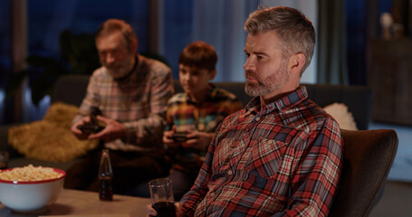 Caucasian father cheering up for favorite football team watching Tv in living room. His son and dad playing console videogames together.