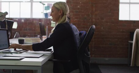 Caucasian businesswoman sitting at desk using computer passing document to female colleague