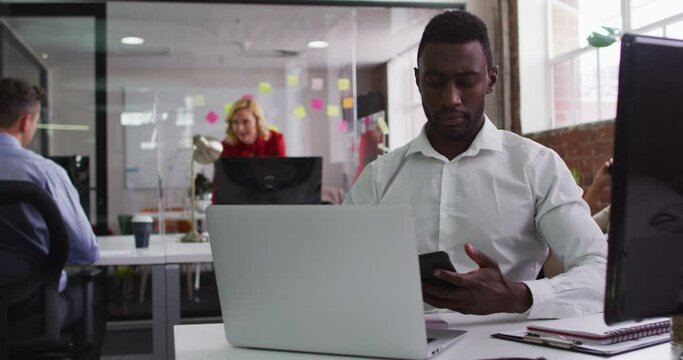 African american businessman sitting at desk talking on smartphone using laptop