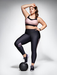 Obraz premium Sporty girl resting after workout with medicine ball. Photo of model with curvy figure in fashionable sportswear on grey background. Sports motivation and healthy lifestyle