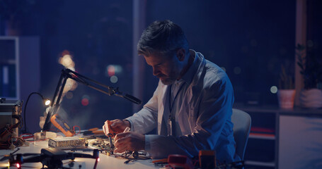 Bearded adult engineer in lab white coat sitting by desk soldering iron circuit board fixing drone device. Technical repair, research office. Engineering.