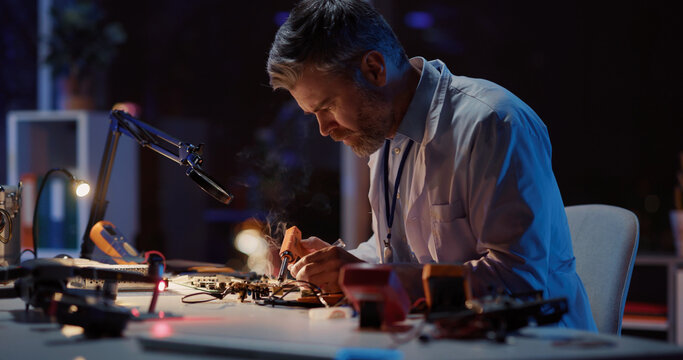 Caucasian adult engineer technician in lab coat sitting inside office soldering circuit board of broken drone using modern equipment working at night.