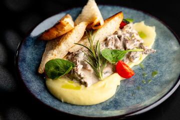 Meat in a creamy sauce with mashed potatoes and bread toast, painted with spinach leaves and cherry tomatoes, on a gray plate on a black background.