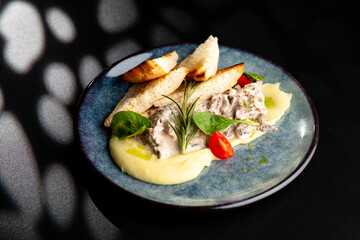 Meat in a creamy sauce with mashed potatoes and bread toast, painted with spinach leaves and cherry tomatoes, on a gray plate on a black background.