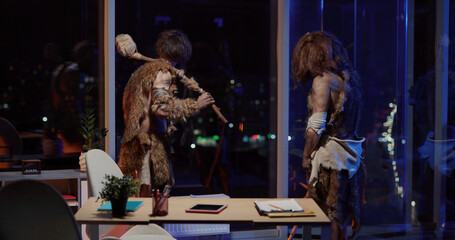 Primeval couple of two savages with prehistoric tools standing in corporate modern office looking...