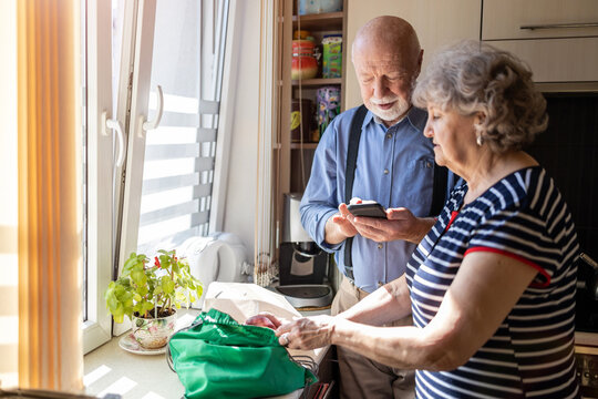 Senior couple using a smartphone while unpacking groceries in the kitchen at home