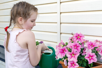 Lovely little girl outdoor takes care of flowers by watering it by green watering can. Activities with children outdoors. Summer, spring, gardening concept. Copy space.