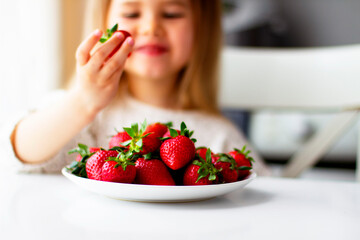 Cute little girl eating fresh strawberry in the kitchen. Healthy vitamin snack for kids. Ripe fresh berries. Harvest season. Natural vitamins .