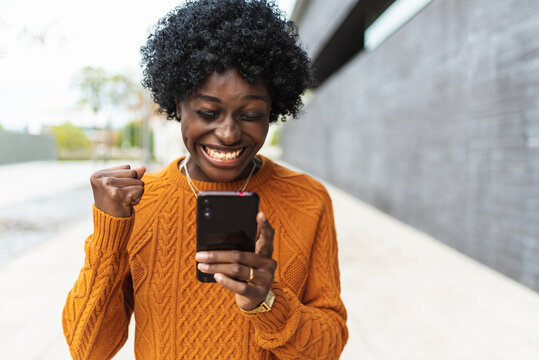 Image of a happy and emotional African American woman posing isolated on out of focus background using cell phone making a winning gesture.