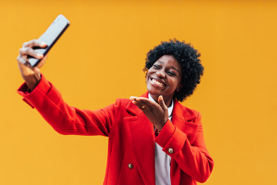 African American brunette woman taking a selfie and smiling on a yellow background outdoors.