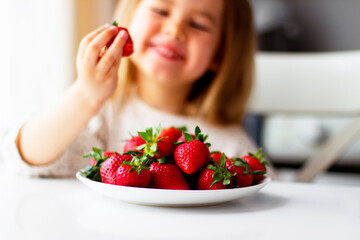 Cute little girl eating fresh strawberry in the kitchen. Healthy vitamin snack for kids. Ripe fresh berries. Harvest season. Natural vitamins .