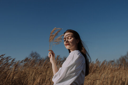 Smiling young woman in white blouse standing in field and holding braches of dry pampas grass in front of sky. Style and fashion. Girl in casual outfit with closed eyes enjoying sunlight. Golden hour