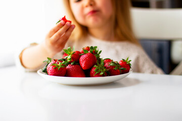 Cute little girl eating fresh strawberry in the kitchen. Healthy vitamin snack for kids. Ripe fresh berries. Harvest season. Natural vitamins .