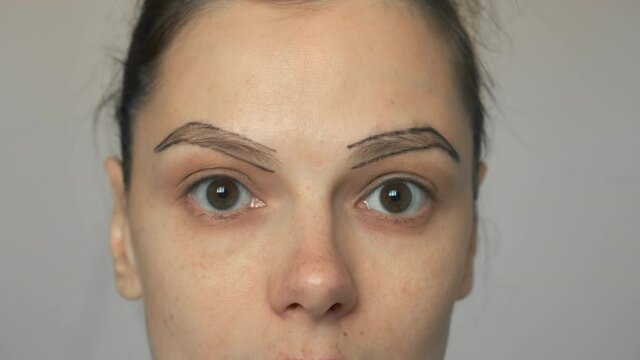 Young woman with painted contours of eyebrows before painting them, closeup face. She is looking at mirror. Preparing to eyebrow paint beauty procedure for herself at home. Female without makeup.