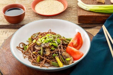 Soba with mushrooms, green beans and sesame seeds in a plate on a stand on a wooden table next to sticks and soy sauce and sesame seeds.