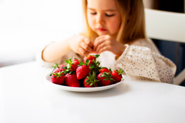 Cute little girl eating fresh strawberry in the kitchen. Healthy vitamin snack for kids. Ripe fresh berries. Harvest season. Natural vitamins .