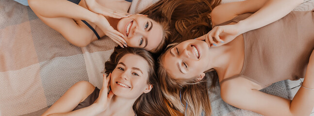 Women rest and have fun. Girlfriends laugh at home lying on the floor on pillows. Tree girls make homemade face and hair beauty masks. Women take care of youthful skin. - obrazy, fototapety, plakaty