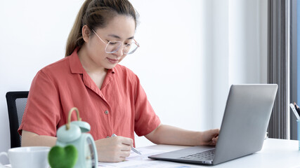 Fototapeta premium Asian business woman in a bright t-shirt is working at home, Stay home, New normal, Covid-19 coronavirus, Social distancing, Coming up with ideas for work, Writing information in a notebook.