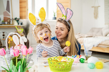 Beautiful happy son and mother in headbands with bunny ears decorate eggs with a brush and bright paints, prepare to celebrate Easter, have fun at home