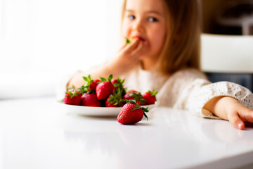 Cute little girl eating fresh strawberry in the kitchen. Healthy vitamin snack for kids. Ripe fresh berries. Harvest season. Natural vitamins .