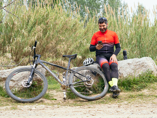 Obraz premium Male mountain biker in nature next to his bike using his mobile phone