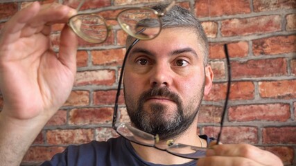 A bearded man tries on different glasses. Short haircut. Blue T-shirt. Poor eyesight. Choice of glasses. Optics. Against the background of a brick wall. Large portrait.