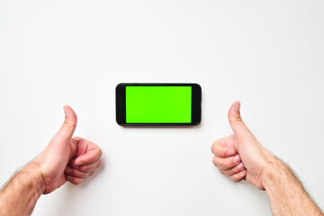 Woman hand shows mobile smartphone with green screen in vertical position isolated on green background with thumbs up finger. Mock up mobile screen blank space