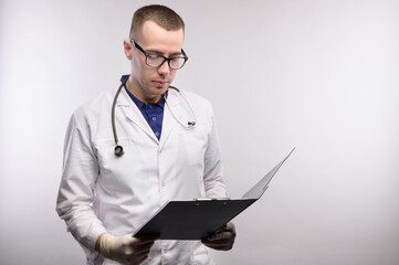 Portrait of an attractive young Caucasian doctor in glasses and a white coat stands with a tablet in his hands smiling and looking at tablet on a gray background. Studio portrait copy space