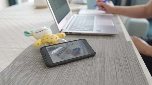 Mother working on laptop using video baby monitor app on phone, woman watching her sleeping baby in real-time on smartphone screen. High quality 4k footage