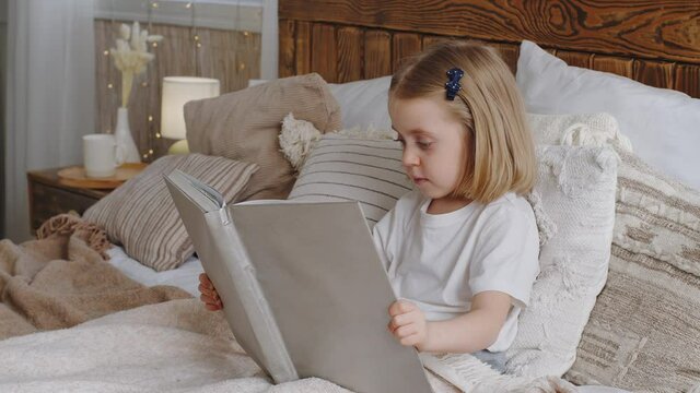 Little cute girl small daughter kid child baby toddler preschooler sits in bed in cozy bedroom interesting reads book fairy tale looks at pictures in grey photo album before sleeping, home education