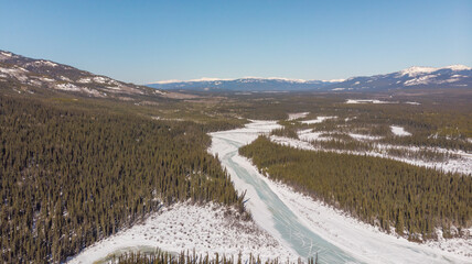 Drone, aerial view of a frozen, thawing river in northern Canada with surrounding wilderness from the Alaska Highway. Taken in April, spring time with perfect blue sky. Desktop background view. 
