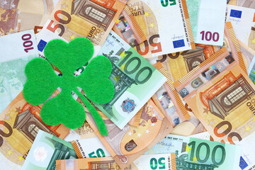Green felt four leaf clover on surface covered with 50 and 100 euro banknotes, on the left side. Financial, bank, money, economy, business concept background. Lucky talisman. Saint patrick's day.