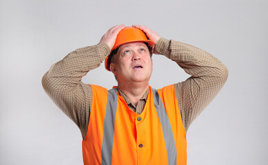 contruction worker clutching his head in helmet on grey studio background looking up, foreman shocked